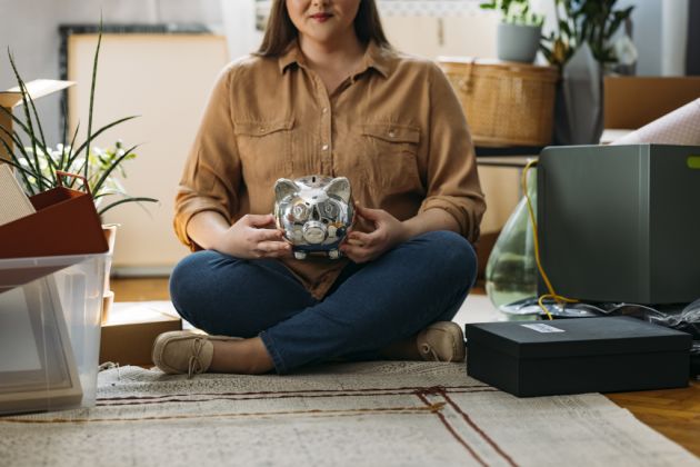 Unrecognizable Caucasian plus size woman sitting on the floor among boxes with personal belongings, holding a piggy bank while facing camera ( horizontal, cropped, copy space)