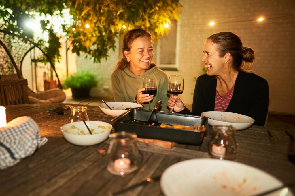 Two laughing female friends toasting together with glasses of wine after a candlelit dinner outside on their patio in the evening