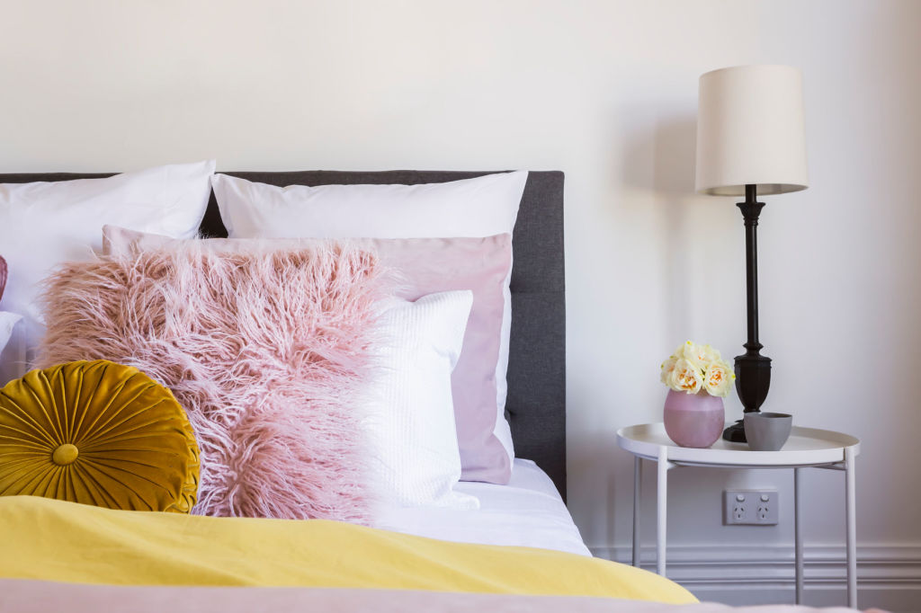 A bedside table with flowers, vintage lamp and pink and yellow bed linen Photo: Stocksy