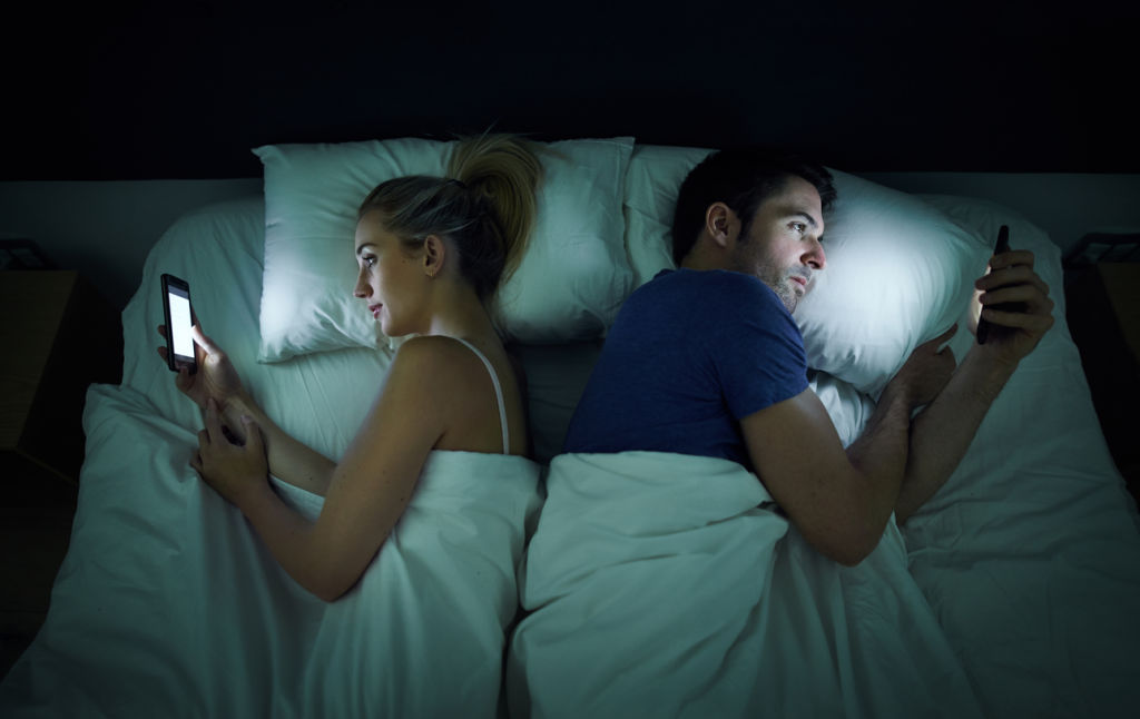 A young couple use their cellphones in bed at night. Photo: A.J.Watt (iStock)