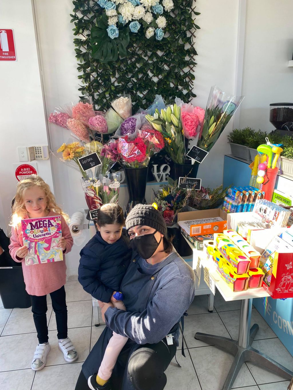 Chiswick art contest winner Ellie Harrison, five, holding her colouring book prize, with Samantha Touma, Chiswick Convenience shop owner, with Ellie's friend Emma Manto, also five. Photo: Supplied