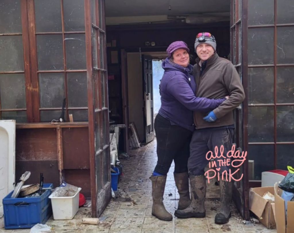 Man and woman in jackets hug in front of rundown house.